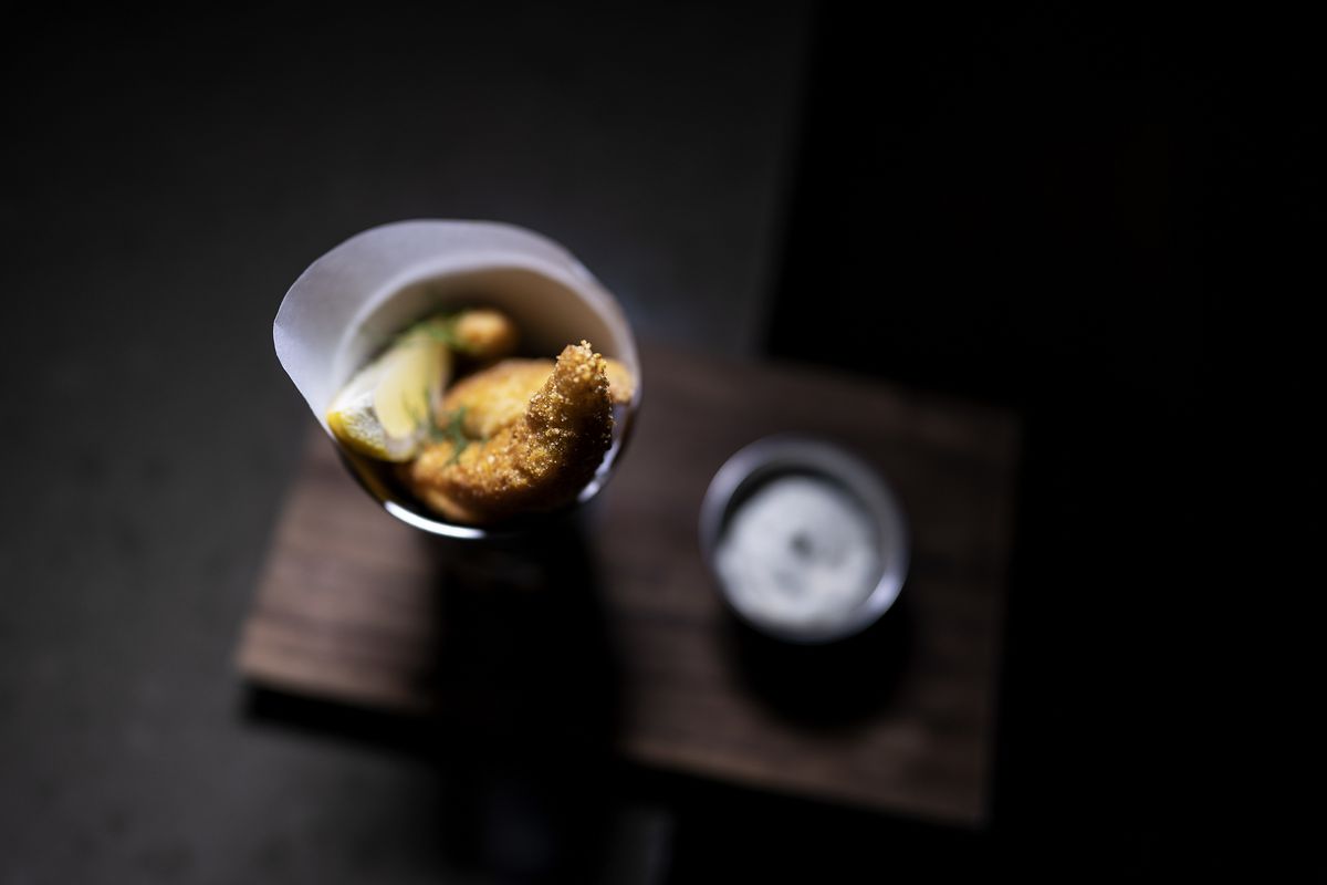 Fried fish strips in a metal cup, served on a board with a side of white, creamy sauce