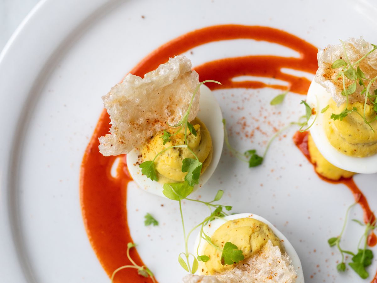 Three deviled eggs on a plate decorated with a swirl of red liquid. The eggs are topped with pea shoots and pork rinds.