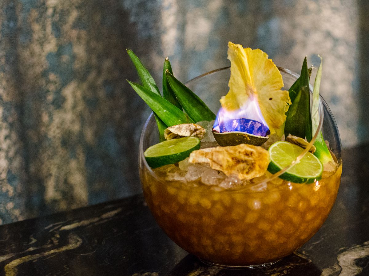A spherical glass punch bowl is filled with an icy cocktail garnished with a lime skin on fire, fresh limes, banana leaves, and more.