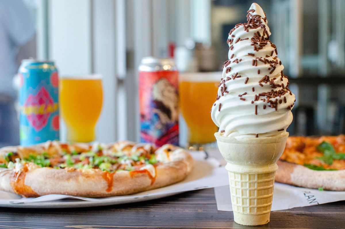 A cone of vanilla soft serve with chocolate sprinkles sits on a restaurant table. Blurred in the background, two pizzas and two colorful cans of beer are visible.
