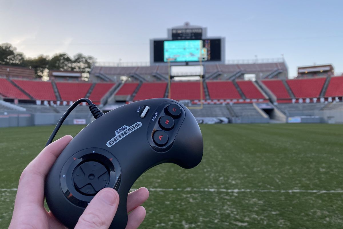 a hand holds a Sega Genesis gamepad with a large stadium video board far in the distance