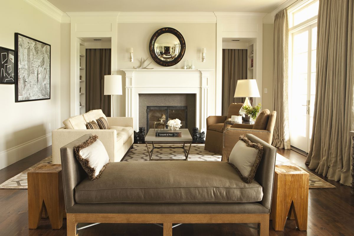 A living area with couches, arm chairs, a fireplace, works of art, a mirror, lamps, and an area rug. The walls are painted with a light grey beige color. 