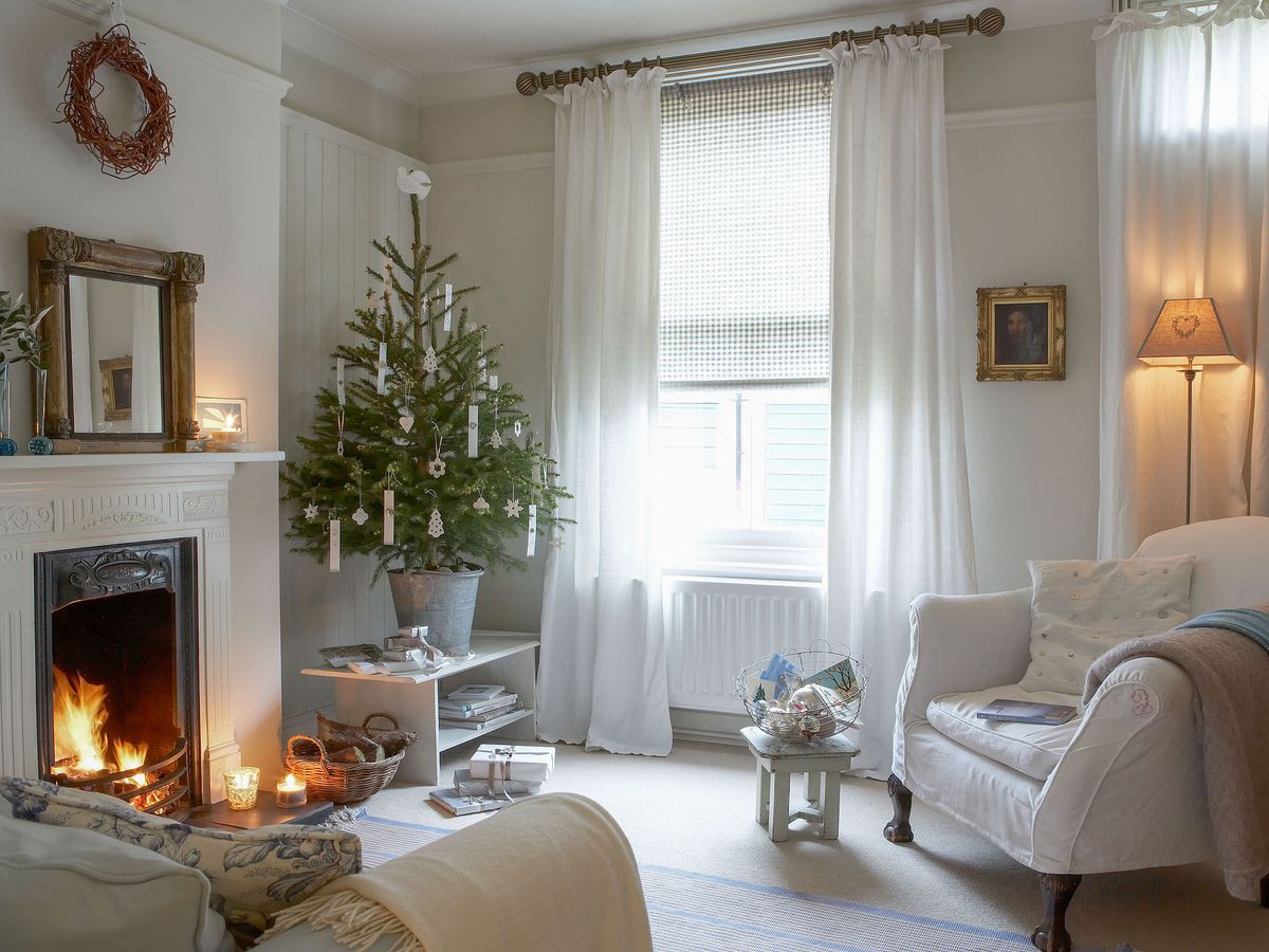A white living room with a small Christmas tree, fire, and flowing drapes on the window. 