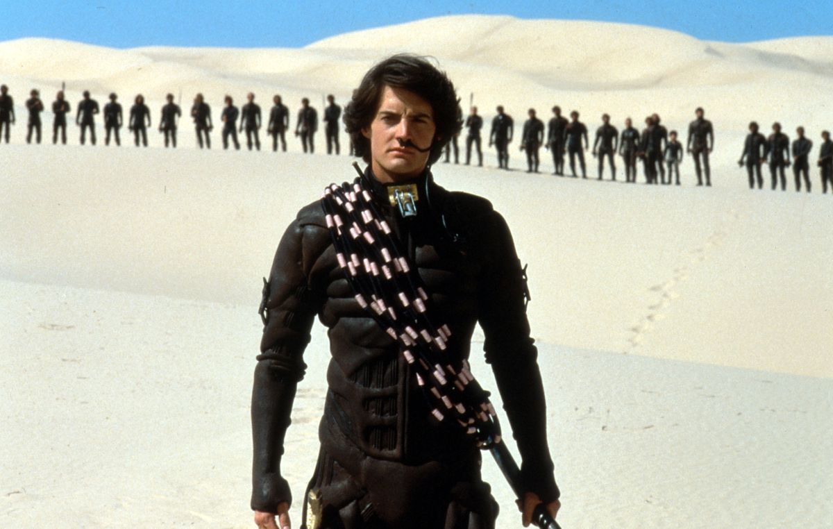 A white young man in a black armor outfit stands against the white sands of a desert, with a line of people in black suits behind him in the desert.