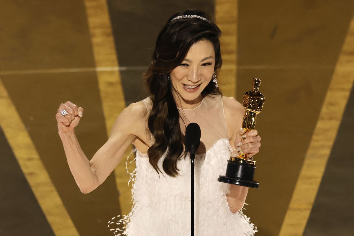 Michelle Yeoh standing at a microphone, holding an Oscar statuette in one hand and making a fist with the other.