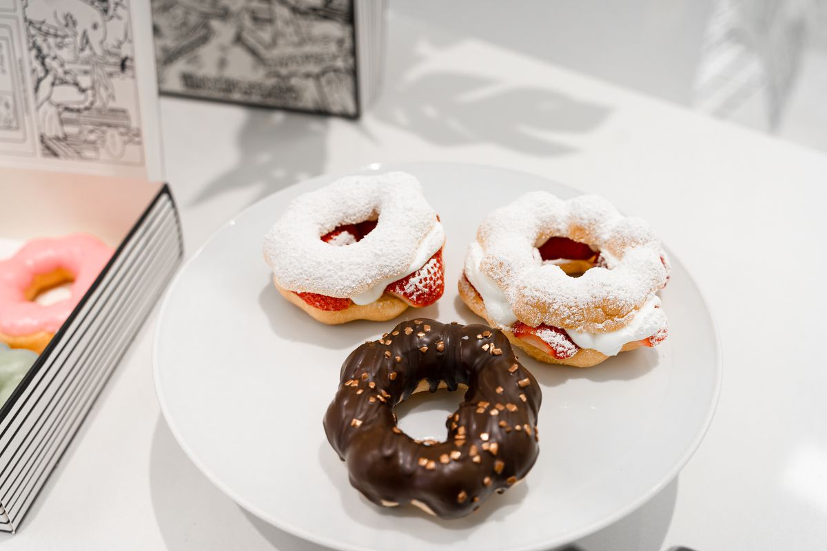 Three pon de ring doughnuts on a plate. Two are covered with white powdered sugar and sandwich sliced strawberries. The third is covered in chocolate.