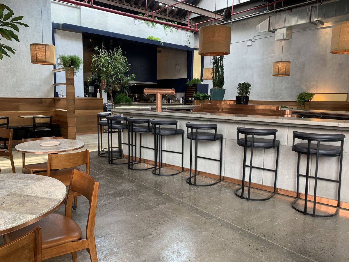 A high-ceilinged, industrial dining room with grey bar stools pulled up to a counter, hanging light fixtures, and plants strewn throughout the space