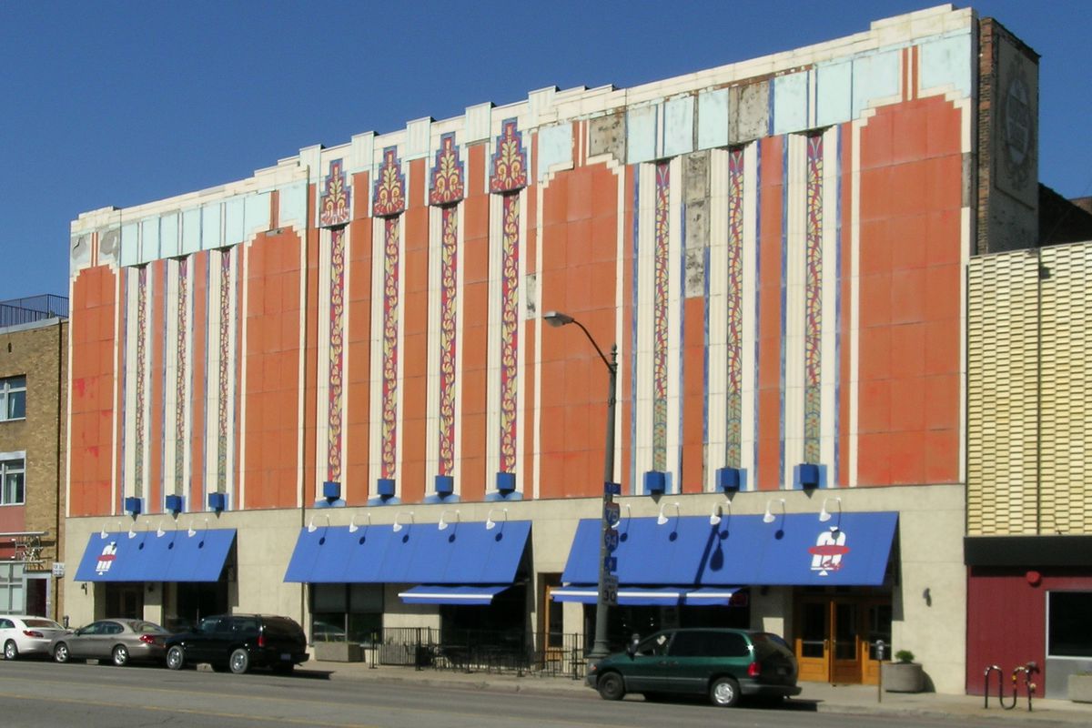 Facade of a several-story Art Deco building with orange paint and vertical strikes of blue and white. There’s also a blue awning over the ground-floor windows.