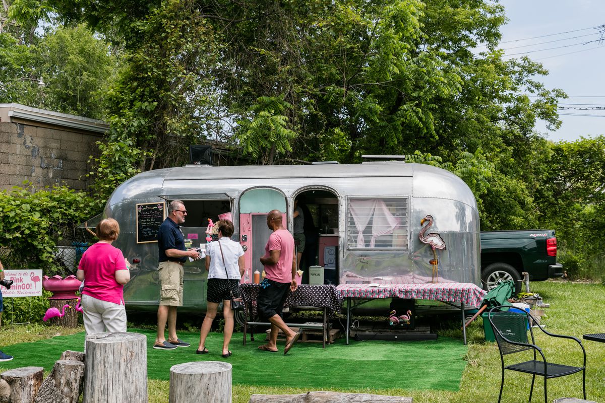 A chrome airstream trailer is parked in a grassy area on a sunny day with a fake grass rug in front. Customers in shorts gather around the trailer.
