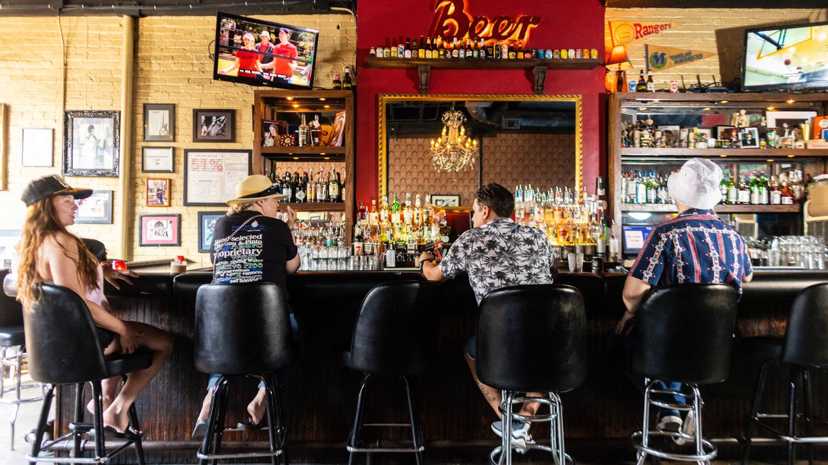 Four people sit at a bar in black faux-leather stools. Behind the bar is a red shelf with a neon sign that reads “beer.”