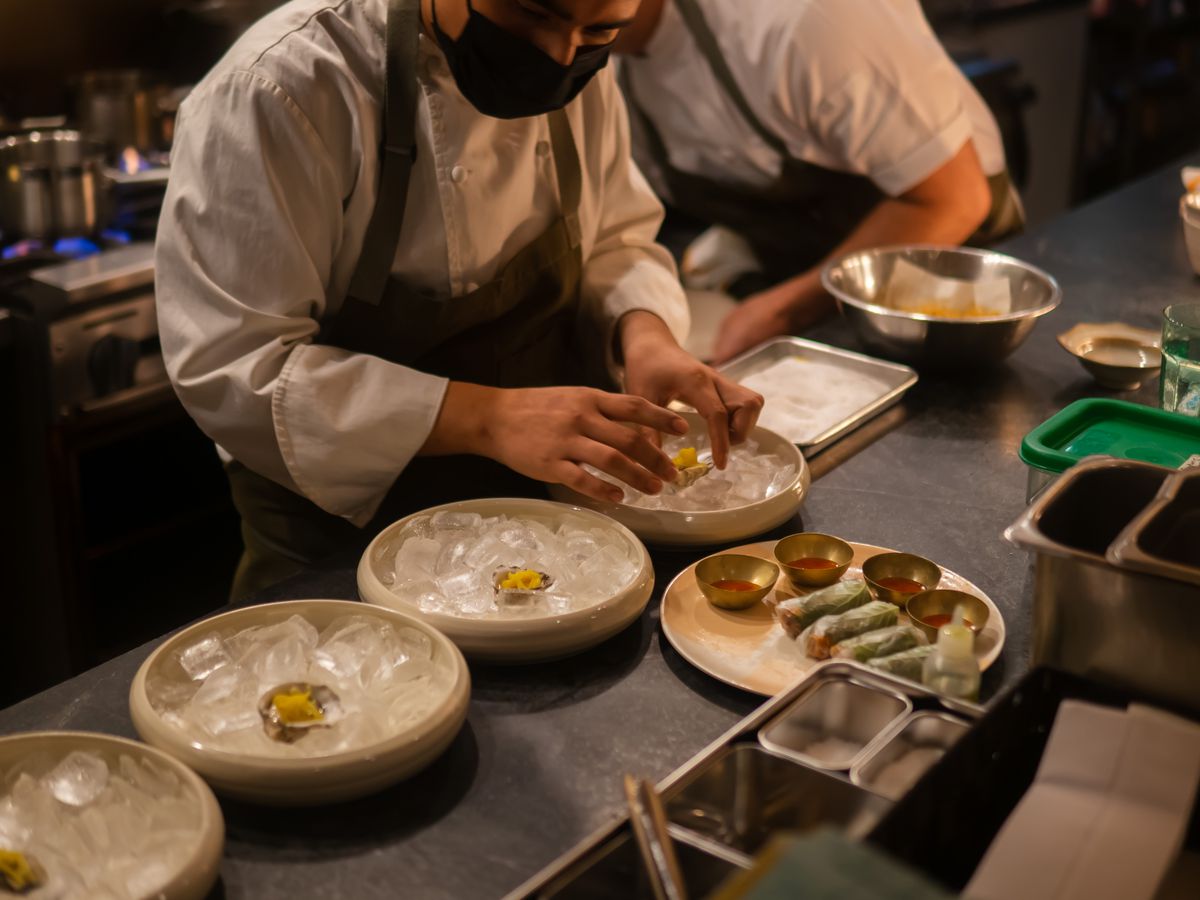 A chef wearing an apron and white shirt, and a black face mask, is plating three ice-filled shallow dishes on a kitchen.