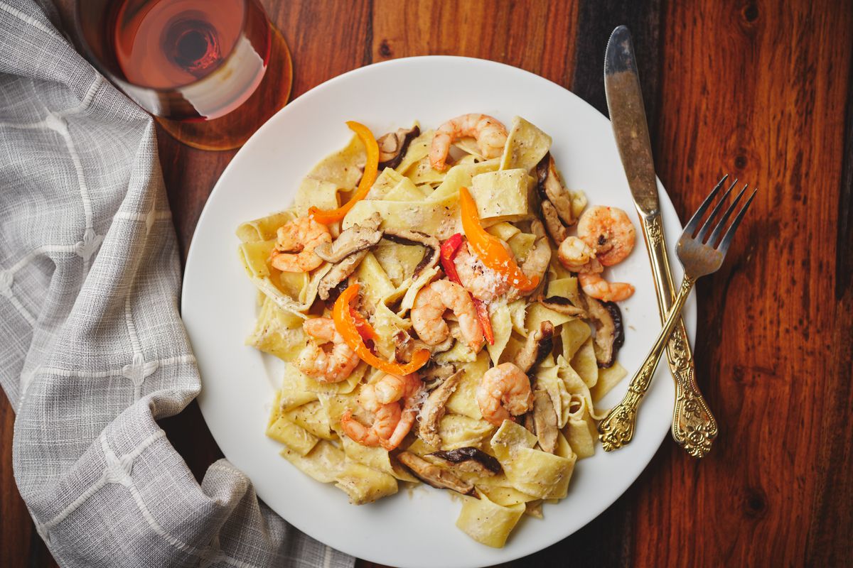 A plate of pasta with shrimp and red peppers sits on a table next to a glass of red wine.