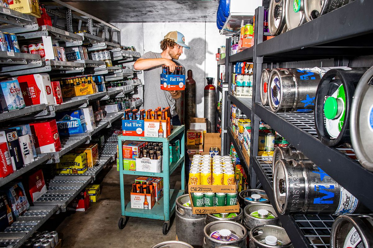 A man with curly hair and a trucker hat holds a case of beer beside rows of beer and kegs.