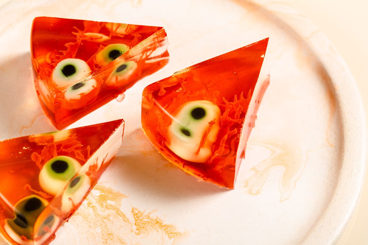 Three slices of a red jelly cake filled with fake eyeballs and booze sit on a cream-colored plate.