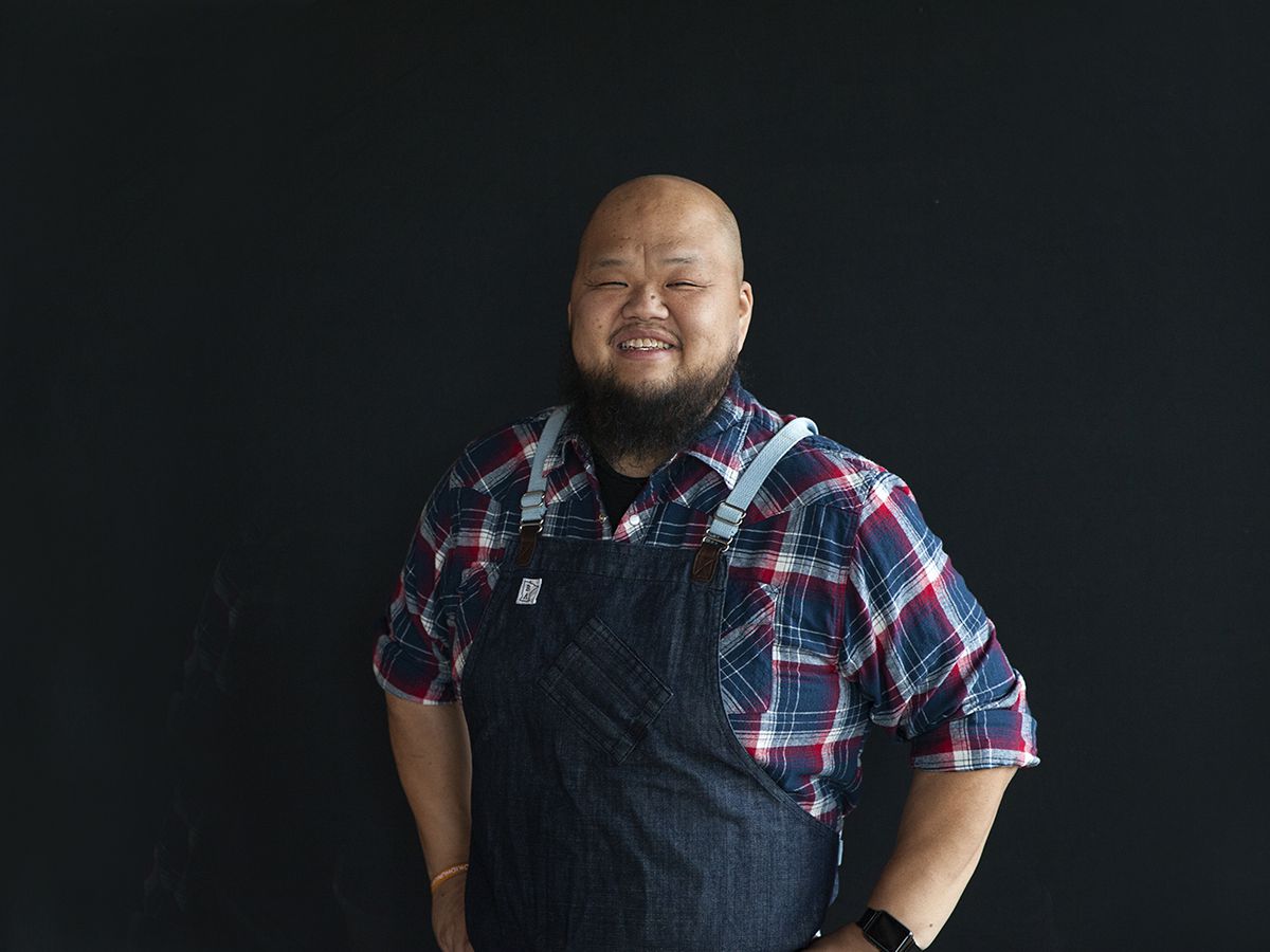 Chef Yia Vang is wearing a plaid shirt and denim apron, smiling, in front of a black background