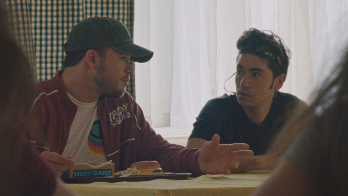Jimmy Tatro sitting at a table wearing a hat while talking to Noah Galvin in Theater Camp