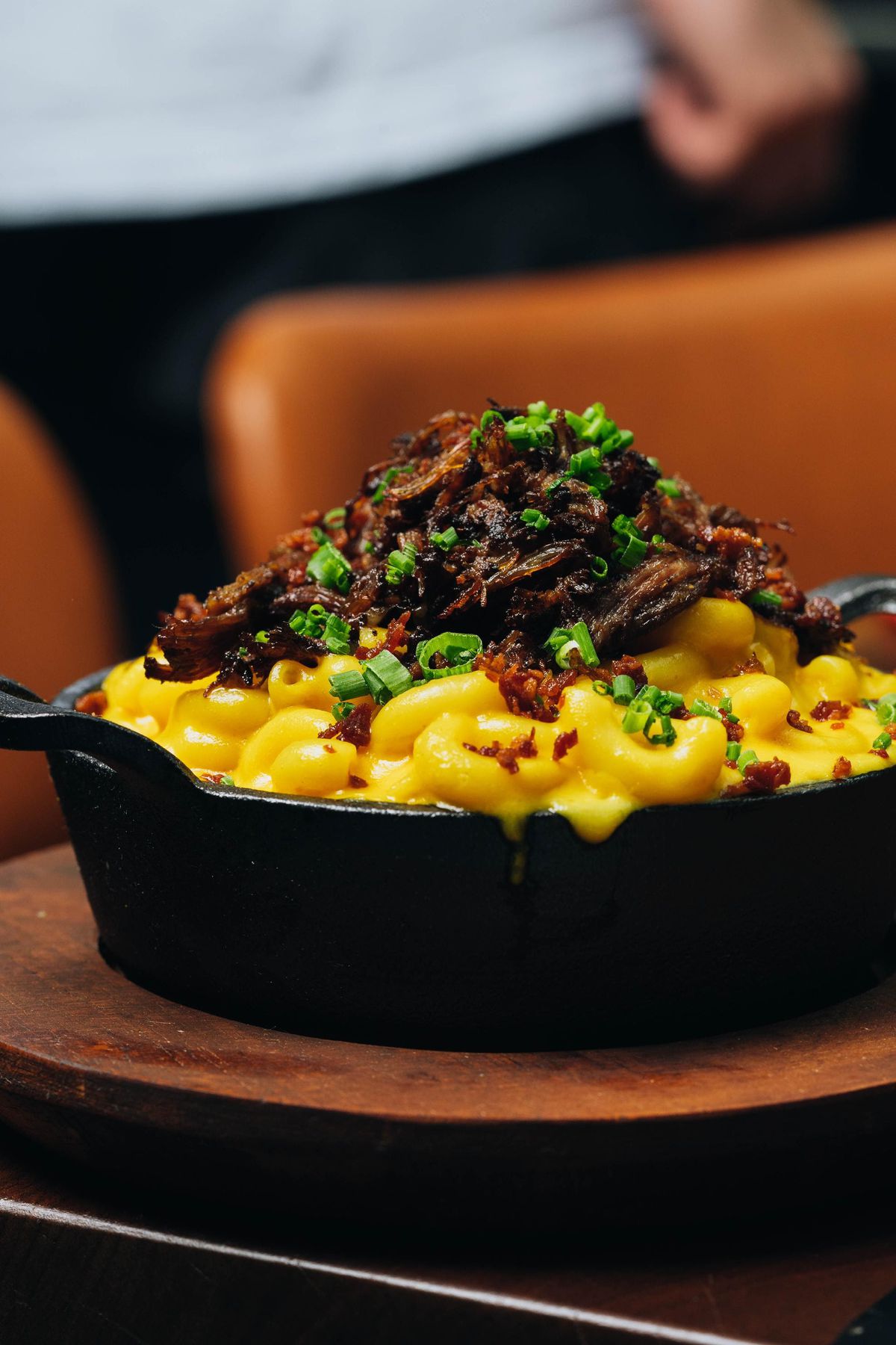 Mac n’ cheese topped with shortrib, bacon, and chives on a brown plate.