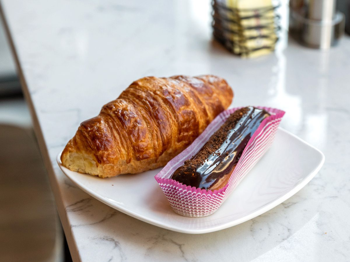 The eclair and croissant are sitting side by side on a plate next to a window. The eclair has glossy chocolate on top and is sitting in a pink and white paper boat.