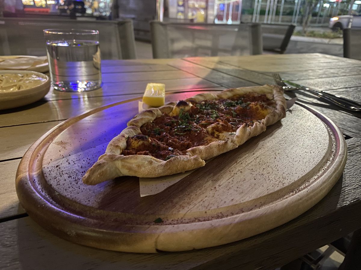 An oblong boat of lamb flatbread with a red center sits diagonally on a wooden platter on an outdoor table
