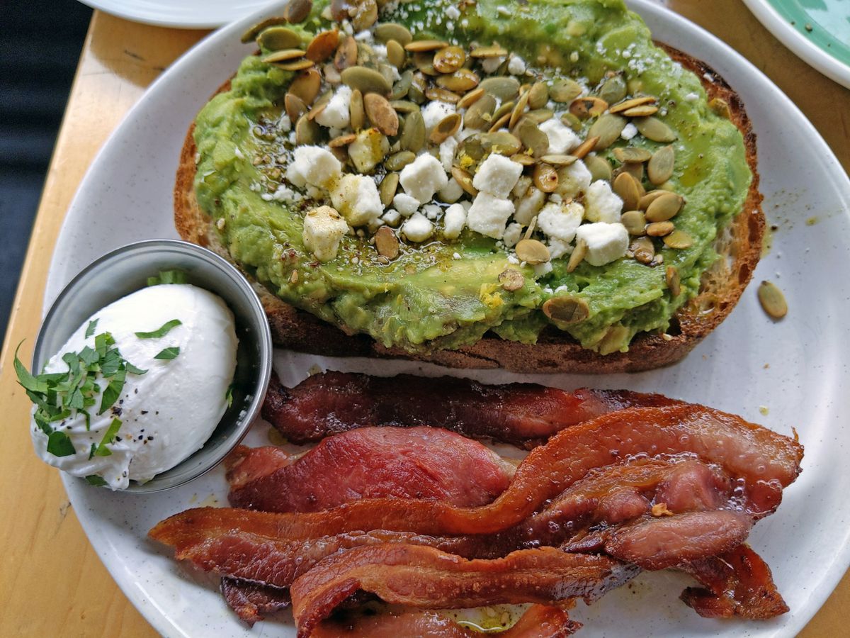 A toast smeared green with seeds on top, and a poached egg in a small bowl and bacon on the side.