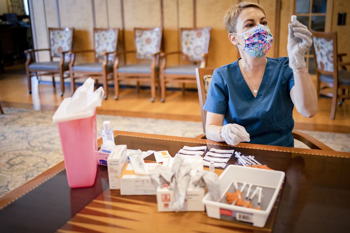 A health care worker sitting at a table with vaccination equipment in front of them holds up a vial and looks at it.