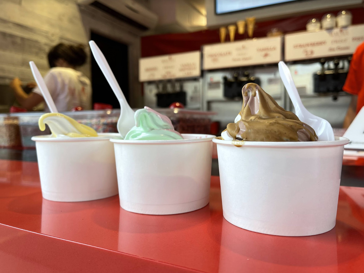 Three cups of soft serve are presented side-by-side in white cups: yellow, green, and brown with a white spoon in each.