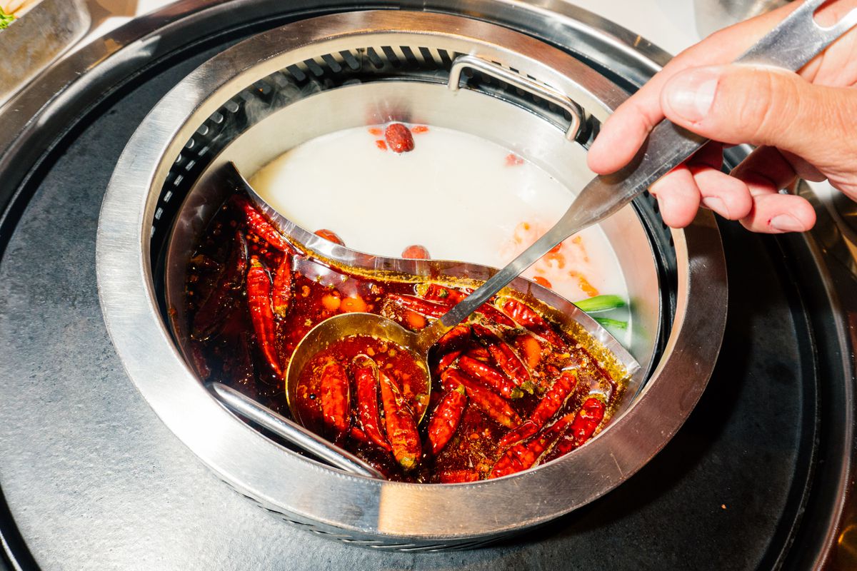 A hand holding a stainless steel spoon ladles red chile peppers in a dark-colored broth.