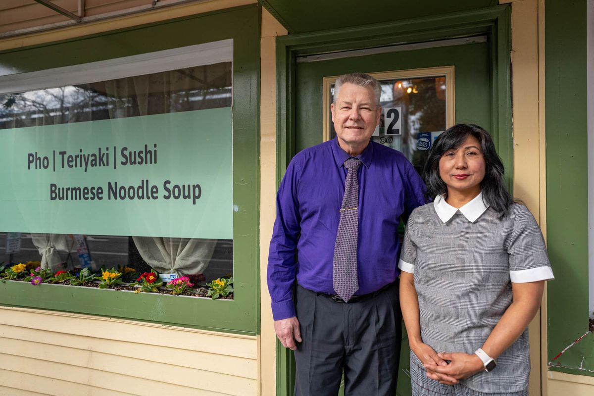 A man in a purple button-down shirt and a tie stands next to a woman with a grey dress in front of a restaurant with “Pho/Teriyaki/Sushi/Burmese Noodle Soup” written on the window.