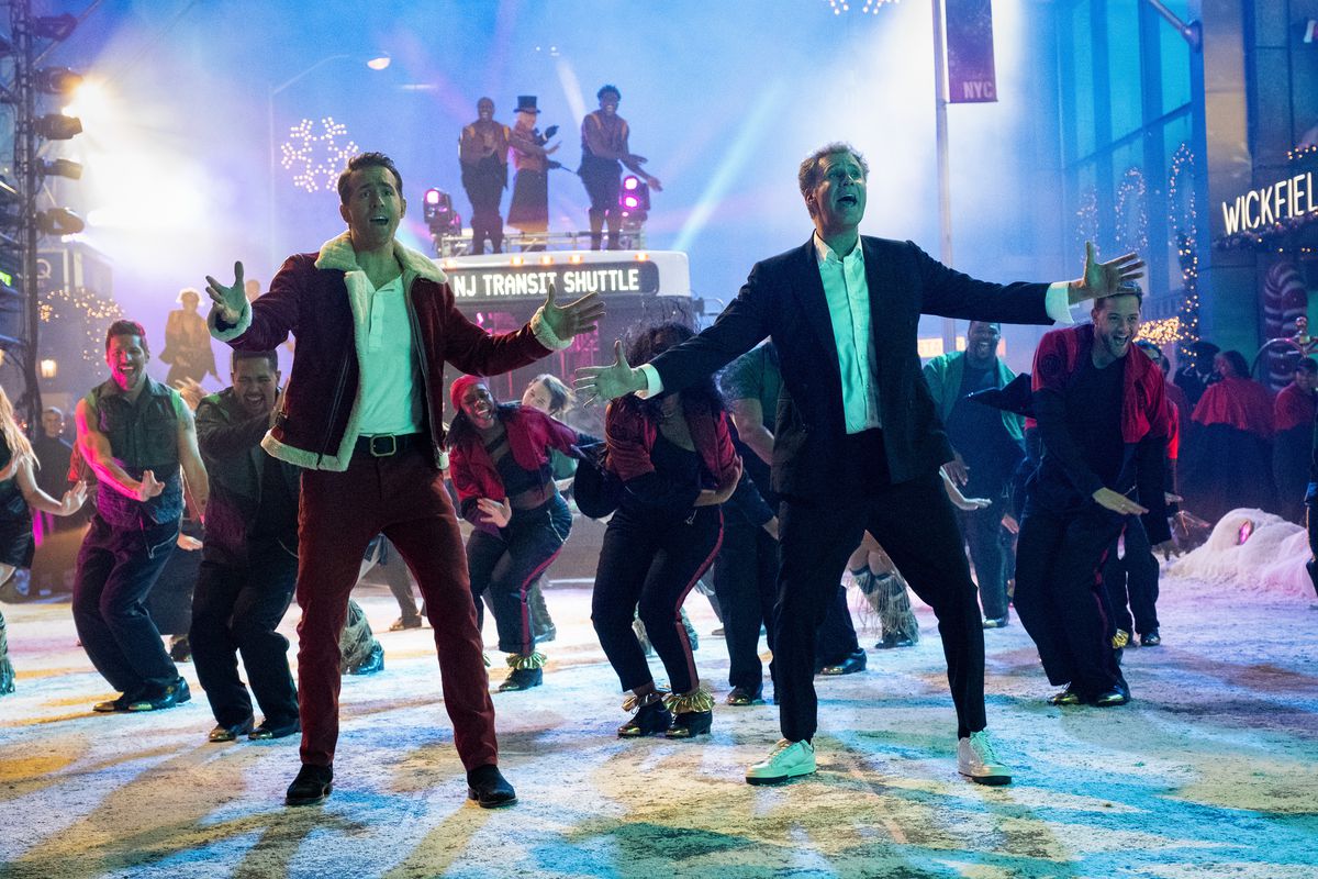 A man in a red and white suit (Ryan Reynolds) and a man in a black and white suit (Will Ferrell) sing alongside one another in from a group of dancers.