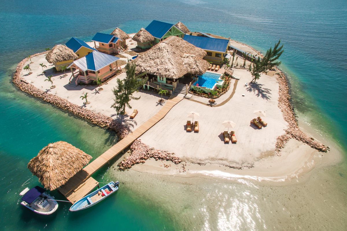 An aerial view of a sandy island with a large thatched roof house surrounded by smaller homes with solar panels.