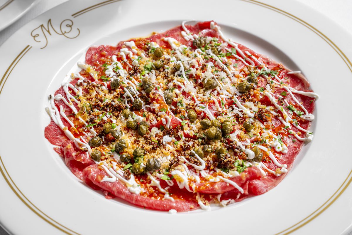 A white plate is almost fully covered with beef carpaccio, which is primarily bright pink and covered with capers, herbs, bread crumbs, and horseradish sauce.