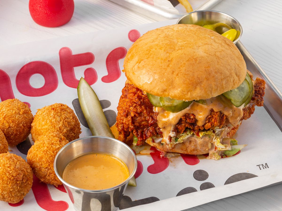 A fried chicken sandwich, leaking sauce and pickles, beside other fried items, on a branded paper-lined tray and squeeze ketchup bottle
