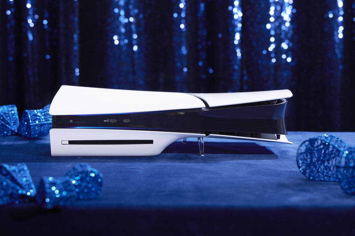 A photograph of Sony’s slim PS5 console laying horizontally on a blue suede surface. There are blue holiday ornaments surrounding the console that are slightly out of focus, as well as a sparkling blue curtain behind the console.