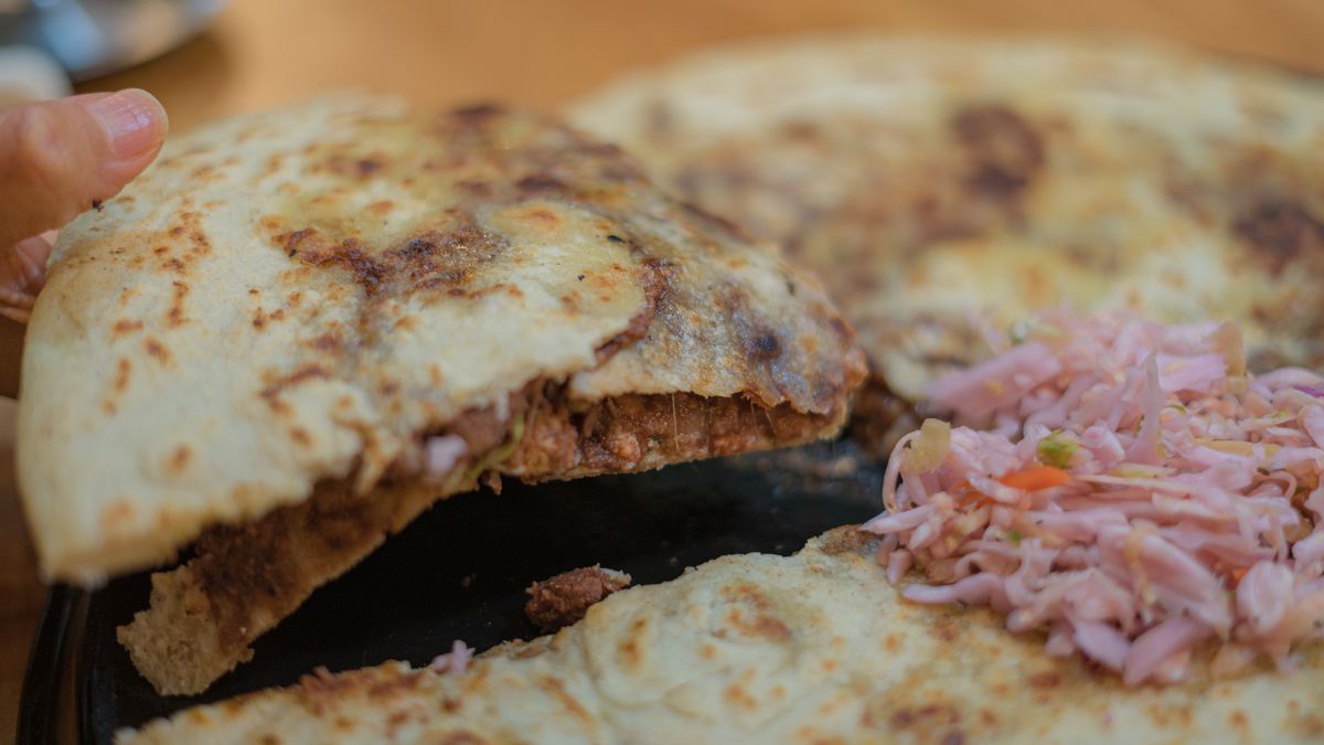 A large pupusa with a golden brown crust with a quarter cut out of it and lifted up by a hand to show the bean, cheese, and pork inside and some pickled purple cabbage on top.