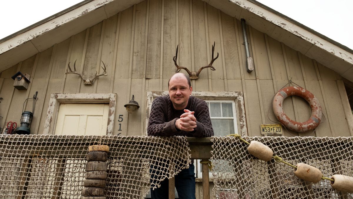 Mike Flanagan stands in front of a wooden house with antlers and a lifesaver on it. He leans against the railing.