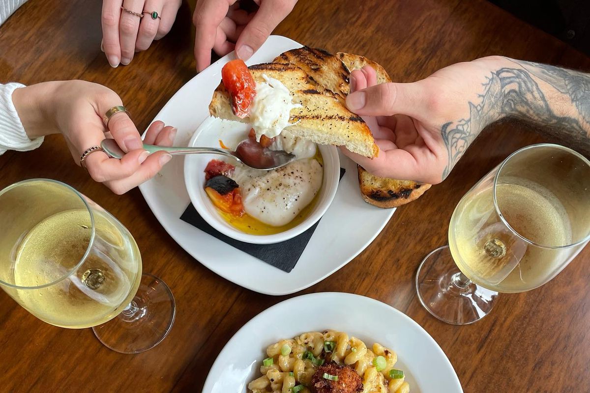 From above shot of two people putting burrata cheese onto a piece of bread with two glasses of white wine and a plate of pasta.