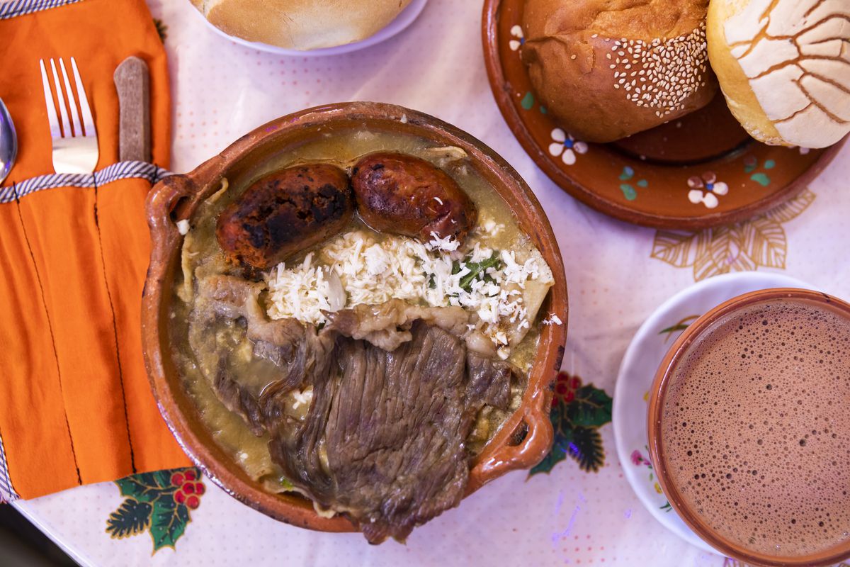 a dish in a clay pot topped with meat with a mug of hot chocolate and some pastries