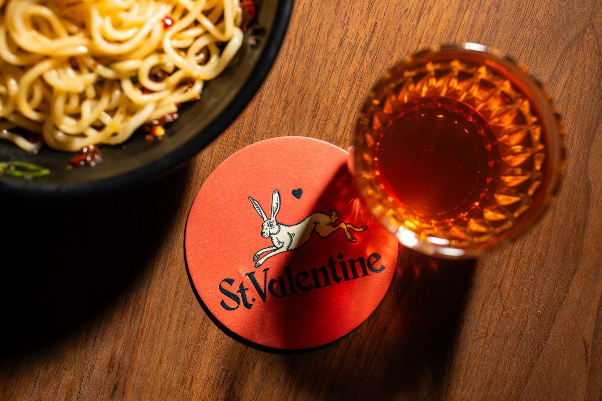 A Sazerac sits on a table with a red coaster next to it that reads Saint Valentine and has a hare on it. A bowl of noodles is visible in the upper right.