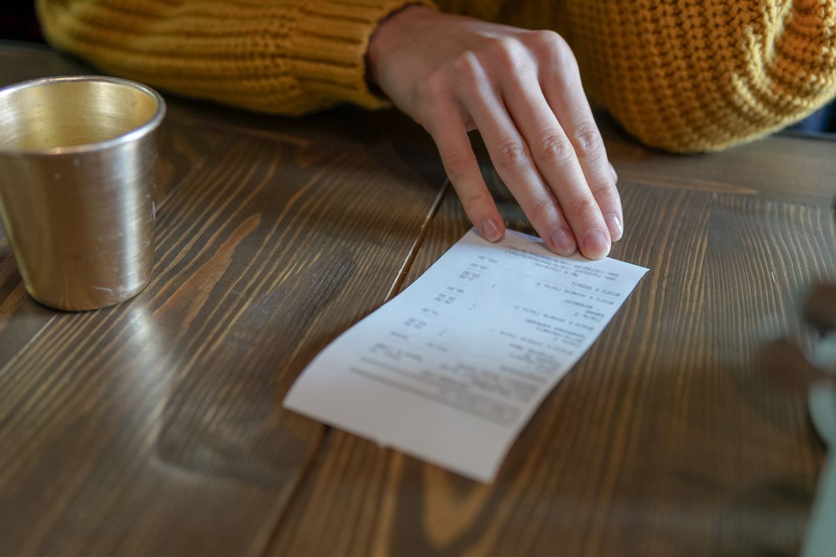 A hand holds a paper receipt on a wooden table with a small metal cup off to the side.
