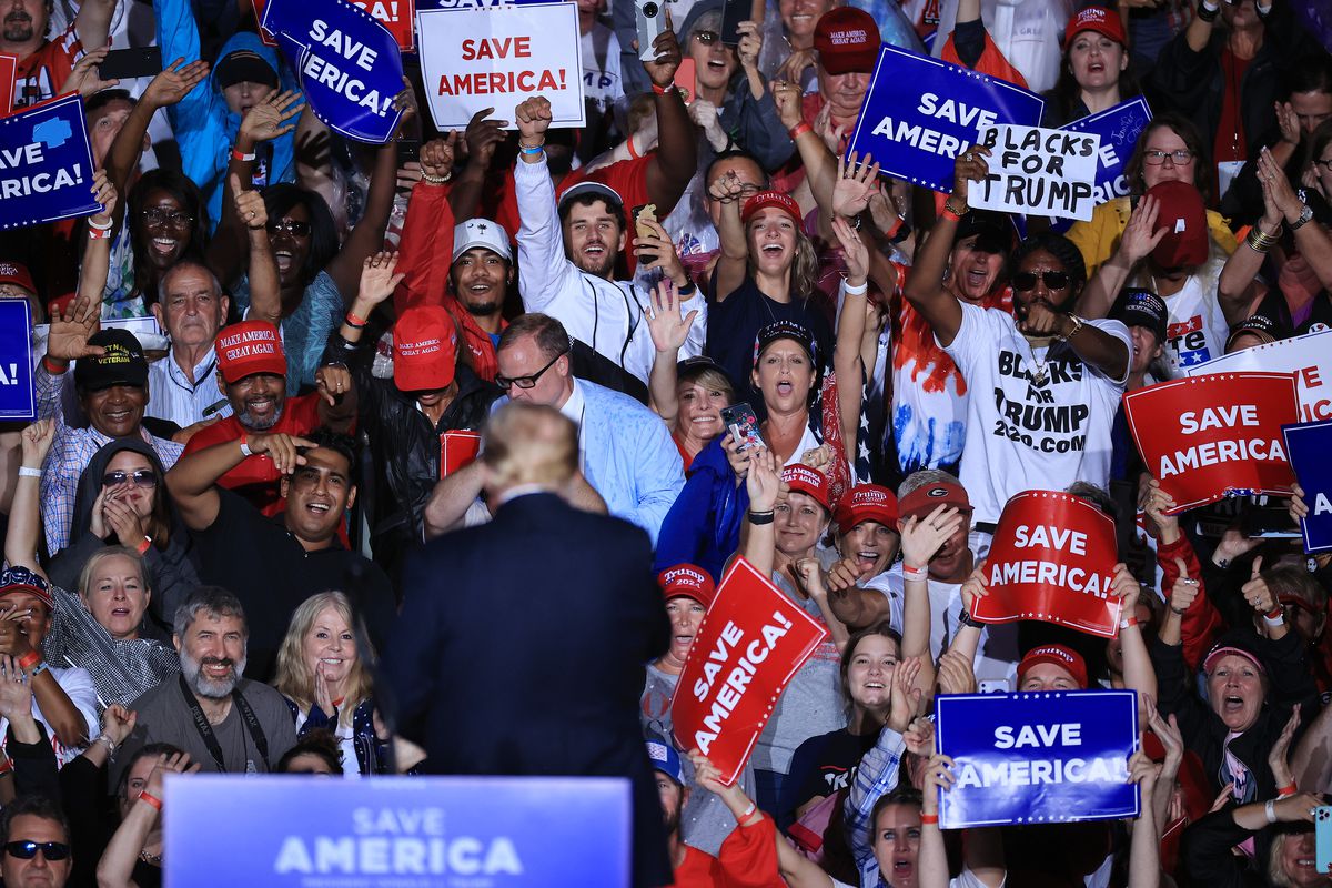 Former President Donald Trump turns from the camera to face the crowd waving “Save America” signs behind him at a rally.