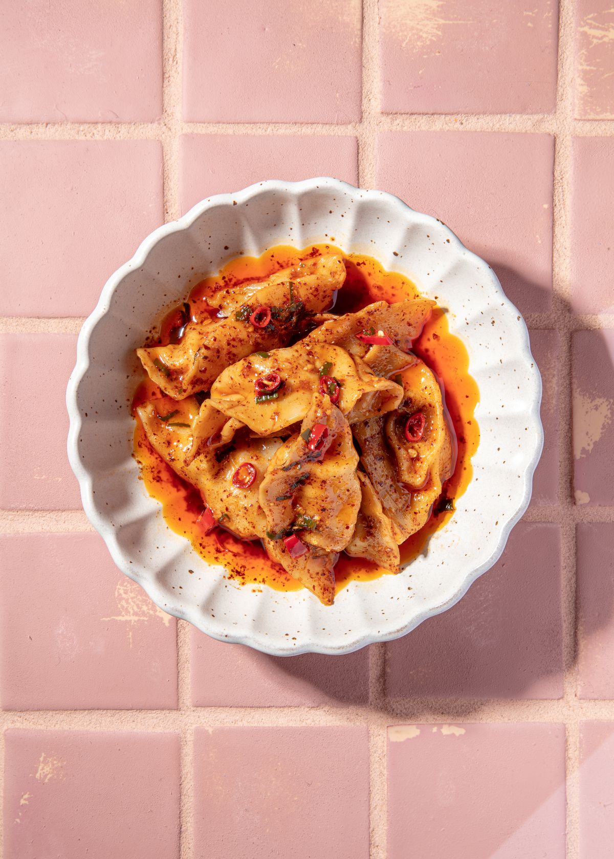 wontons swimming in chili oil in a white dish with a scalloped edge on a pink tile background.