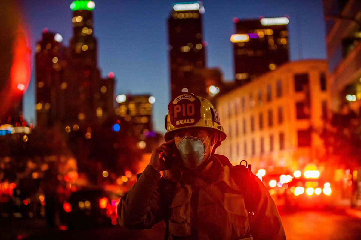 A firefighter holding a cellphone to their ear with a nighttime cityscape and bright lights behind them.