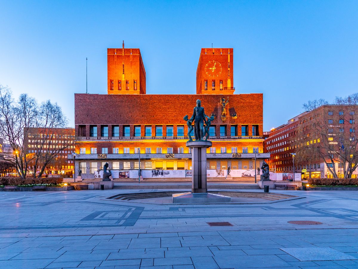 A boxy brick building features two rectangular towers extending from its roof. An open plaza with a sculpture fountain is in the foreground.