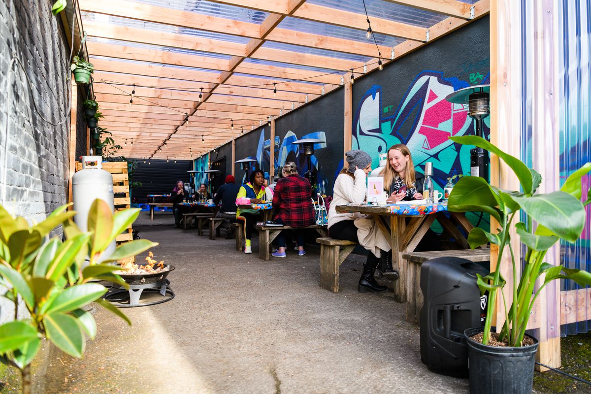 Alongside a graffitied wall, customers sit at picnic tables chatting over cocktails. Heaters stand, unlit, against the tables.