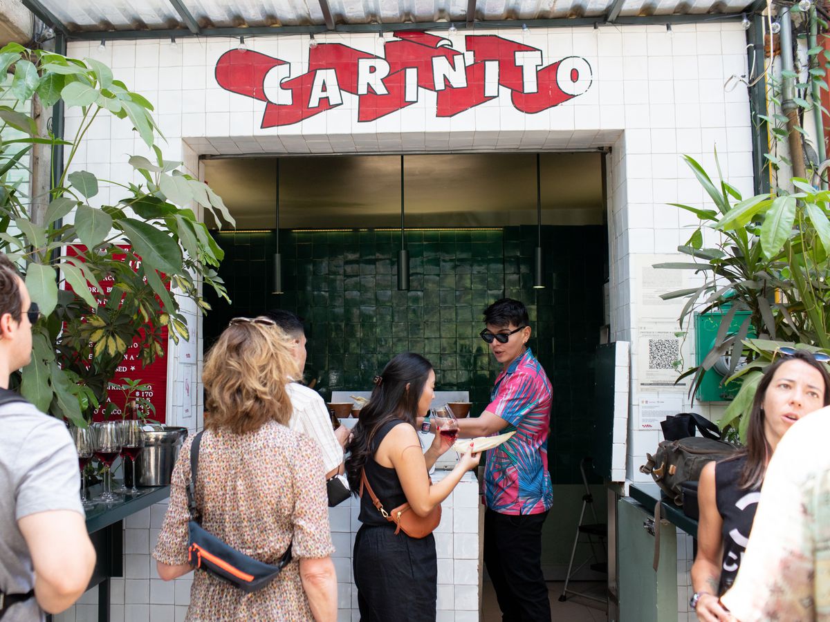 The storefront of a small taco operation with an open kitchen. Customers stand in line while a cook in a floral shirt and sunglasses dispenses food.