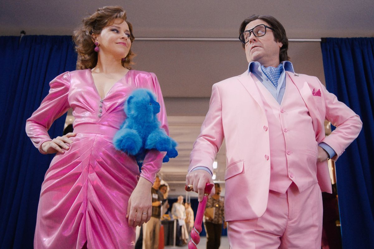 (L-R) Elizabeth Banks and Zach Galifianakis in pink formal attire with a blue beanie baby and a pink cane in The Beanie Bubble.