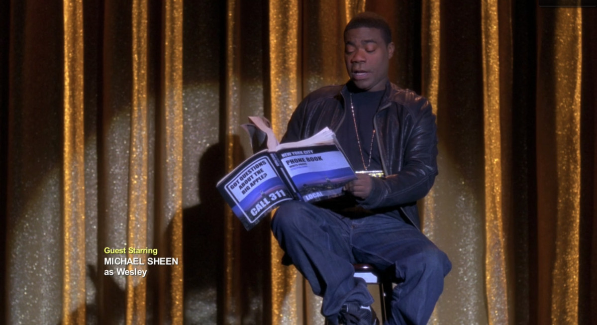 Screeshot of Tracy Jordan in ‘30 Rock’ reading on stage from a phone book