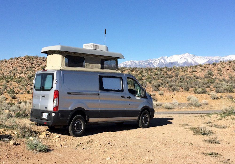 The exterior of a RV camper van, the ModVan CV1. The van is grey and has a white compartment on the roof. It is sitting in a desert with a view of mountains in the distance.