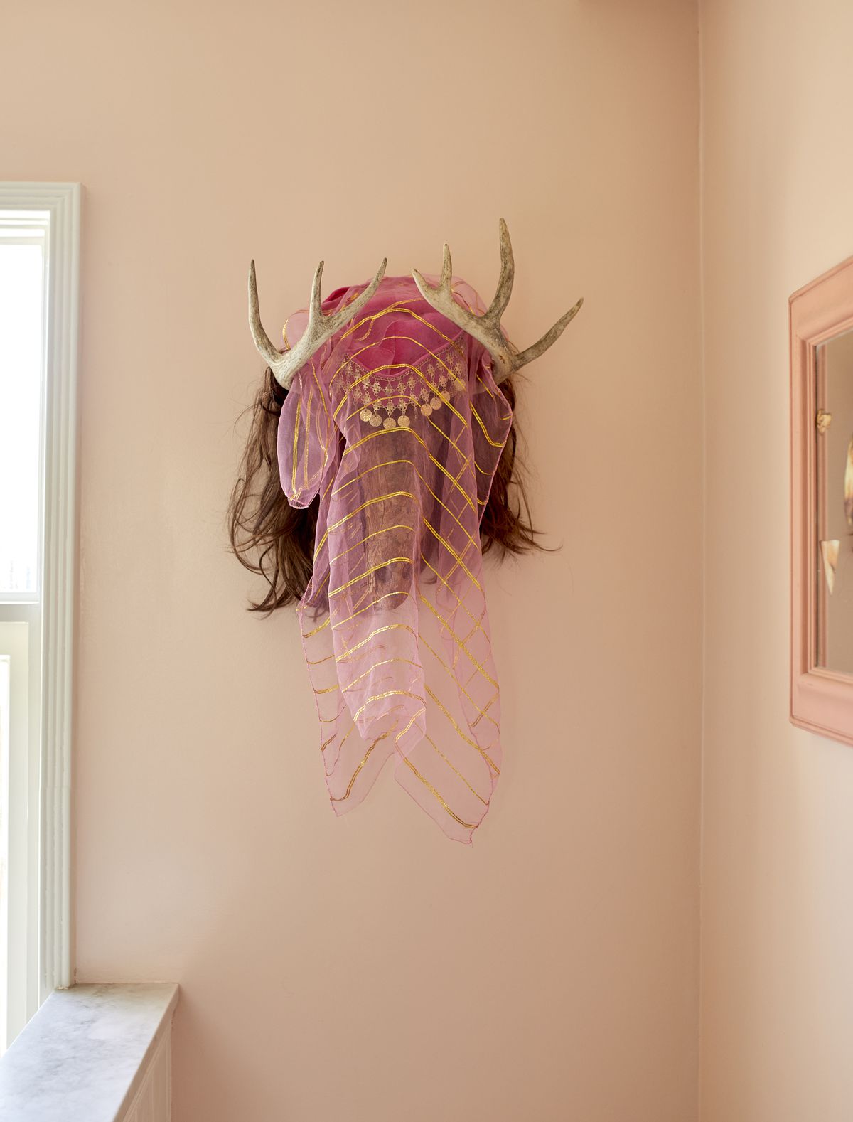 A peach painted wall with a work of art hanging on it. The work of art consists of animal horns, hair, and a pink genie veil. 