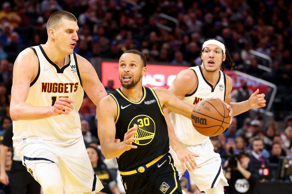 Steph Curry dribbling with his left hand around the defense of Nikola Jokić, with Aaron Gordon in the background. 